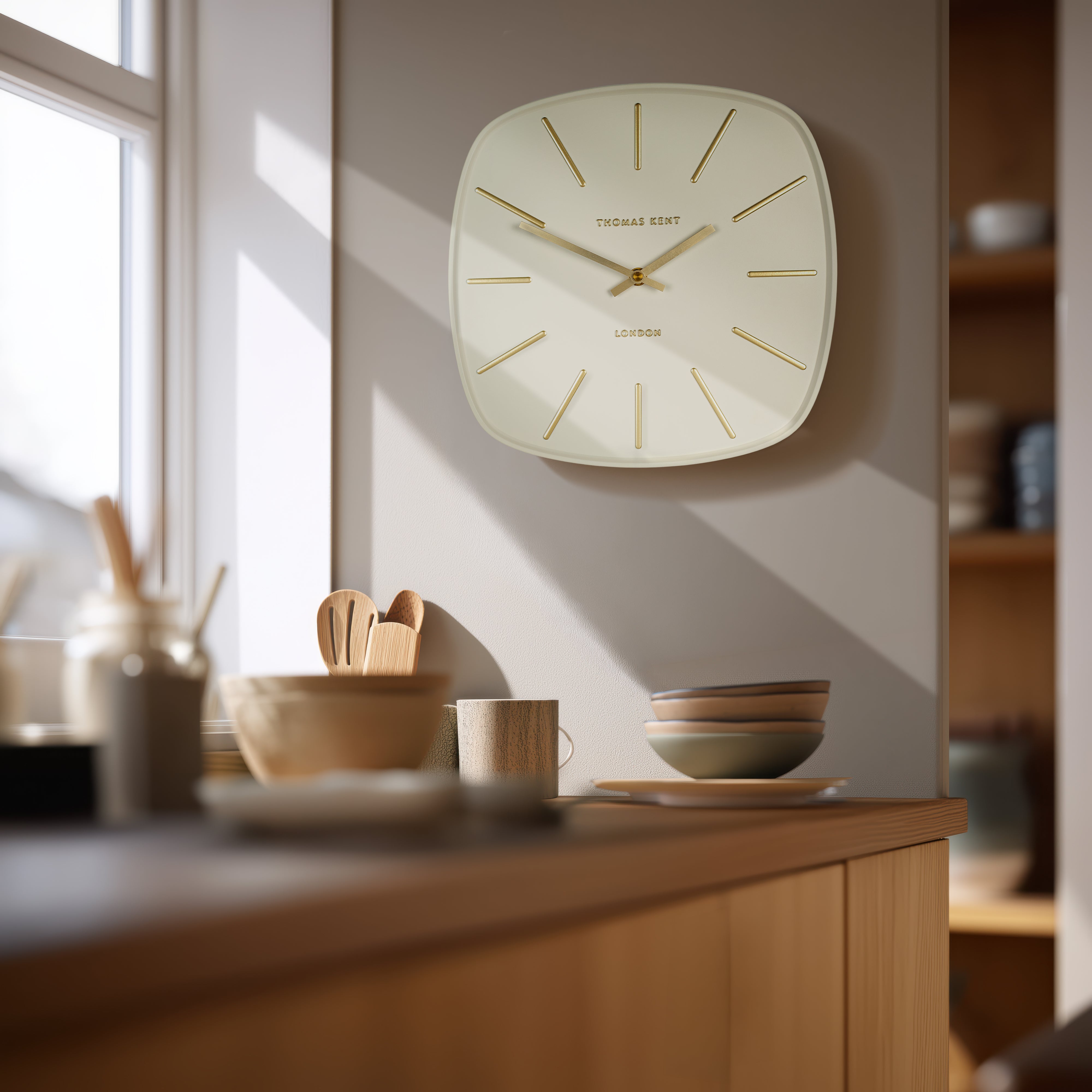 Wall clock on a wall in a kitchen setting with wooden cabinets and utensils.
